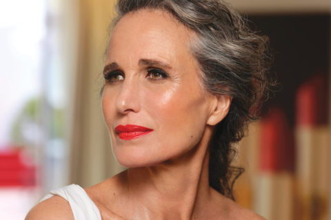 Andie Macdowell close up, wearing red lipstick and other beauty products such as blush and eyeliner. Her hair is back into a loose updo